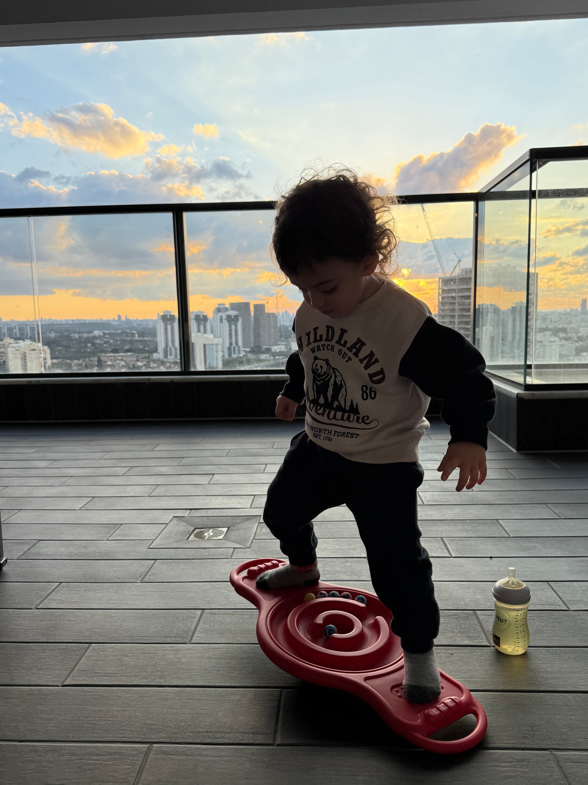 Child playing with a red toy on a wooden floor with a cityscape view through large windows.